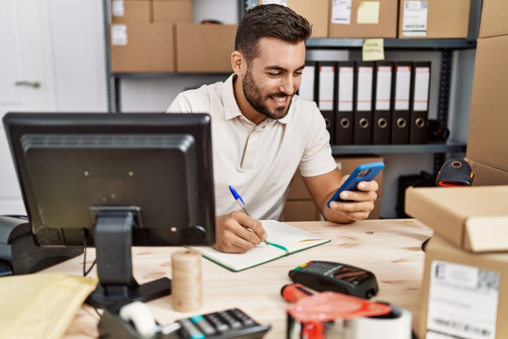 Homem sorrindo e usando smartphone enquanto anota algo em um caderno em uma mesa de escritório com itens de embalagem e pastas ao fundo.